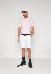 Ljust rosa polo skjorta, vita shorts, svart bälte, svarta strumpor och svarta golfskor. Håller en golfklubba, bär en vit keps.