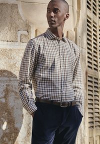 Camicia a quadri marrone e bianca, maniche lunghe, abbottonata, abbinata a pantaloni in corduroy blu navy. Modello in piedi contro uno sfondo di pietra texturizzato.