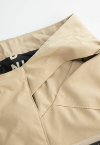 Beżowa podszewka kurtki o gładkiej fakturze, z czarnym wzorzystym wykończeniem i widocznymi detalami ściegów wzdłuż krawędzi.