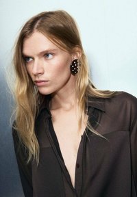 Large, dark brown oval earrings with white dots, paired with a sheer black shirt, showcasing flowing, straight blonde hair and blue eyes.