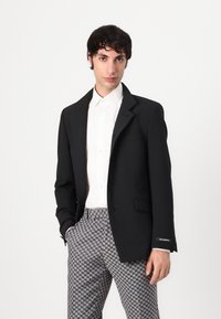 Black blazer with a notched lapel, single button closure, paired with gray houndstooth patterned trousers. White shirt underneath.