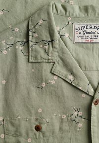Chemise en tissu vert clair avec motif floral rose et branches sombres, bouton en bois et étiquette Superdry cousue.