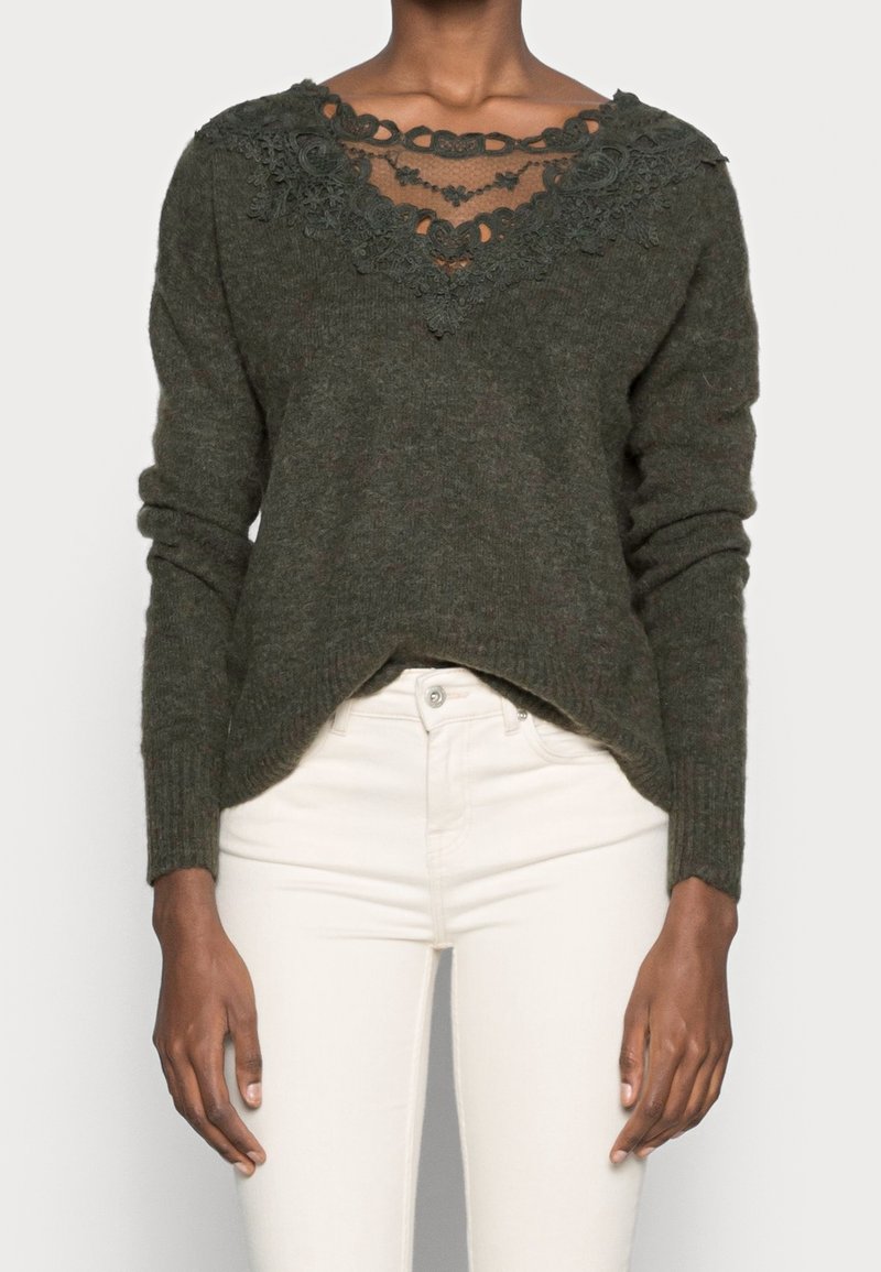 Pull en maille vert foncé avec col en V bordé de dentelle, associé à un pantalon taille haute écru sur une personne debout.