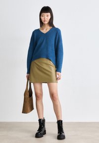 Blå teksturert genser med V-hals, kombinert med en khaki falsk skinn mini-skjørt og svarte snørelests chunky støvler; holder en brun veske.