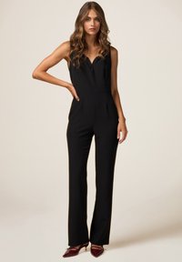 Combinaison noire sans manches avec un décolleté en V, coupe ajustée et jambes droites. Tissu lisse avec un design minimal.