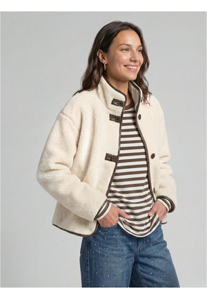 Donna sorridente che indossa una giacca in pile crema sopra una camicia a righe marroni e bianche e jeans blu con piccoli ornamenti di perle.