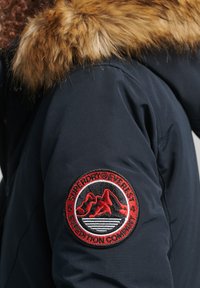 Marineblauwe parka met een afneembare nepbontkap, voorzien van een rood en zwart geborduurd patch met bergen en golven op de linkermouw.