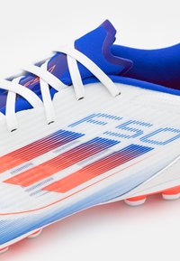 Scarpa da calcio bianca con accenti blu, tomaia testurizzata con strisce diagonali rosse e blu, e lacci bianchi. Suola standard con dettagli arancioni.