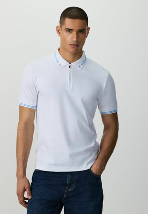 Uomo che indossa una polo bianca a maniche corte con colletto e polsini a righe blu, in piedi con una mano in tasca davanti a uno sfondo semplice.