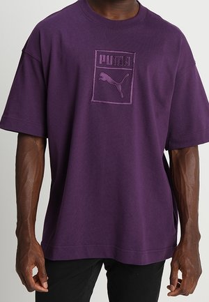 Uomo che indossa una maglietta viola larga con un logo in rilievo di una puma in salto e un testo sul petto, abbinata a pantaloni neri.