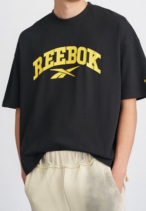Osoba nosi czarną koszulkę Reebok z dużym żółtym logo i jasnobeżowe spodnie z elastycznym pasem, z rękami w kieszeniach.