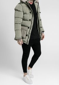 Parka imbottito verde oliva con cappuccio foderato in pelliccia, bottoni neri e tasche frontali, abbinato a pantaloni neri aderenti e sneakers bianche.