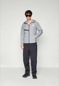 Mann iført grå hettejakke, svarte cargo-bukser, svarte sneakers og store svarte solbriller, stående mot en lys grå bakgrunn.
