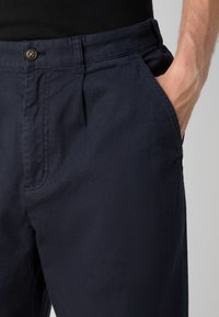 Marineblauwe katoenen broek met een gladde textuur, ritssluiting en zijzakken. Kenmerkt zich door een enkele knop aan de taille en een geplooid ontwerp aan de voorkant.