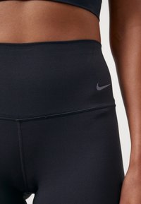 Zbliżenie ciemnoskórej osoby noszącej czarne, sportowe legginsy z wysokim stanem, z małym logo Nike na lewej biodrze.