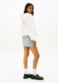 Felpa bianca con cappuccio e cordini, pantaloni shorts in denim azzurro chiaro, calzini bianchi e mocassini neri, che mostrano un design di outfit casual a strati.