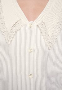 Chemise avec fines rayures noires verticales, trois boutons blancs et un large col en dentelle texturée à motifs floraux.