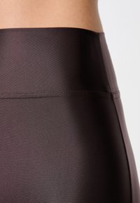 Leggings de tiro alto, de color marrón oscuro, fabricados con un tejido suave y elástico que tiene un ligero brillo, y una cinturilla plana para mayor comodidad y soporte.