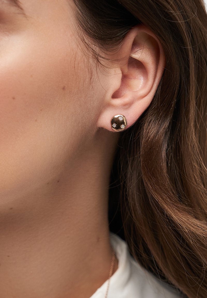 Guldtonade stiftörhängen med en texturerad, rund design och små glittrande accenter. Närbild nära örat för detaljer.