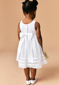 Robe en coton blanche avec un corsage ajusté, une jupe à plusieurs couches avec des rayures en satin horizontales, et un grand nœud dans le dos. Chaussures blanches.