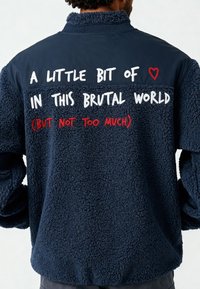 Polaire marine à col montant. Présente un texte blanc et rouge au dos, avec une surface douce et texturée et une coupe décontractée.