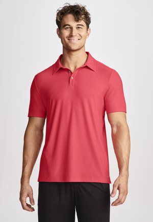 Hombre sonriente con cabello rizado, vistiendo una camisa polo coral de manga corta y pantalones negros, frente a un fondo claro y liso.