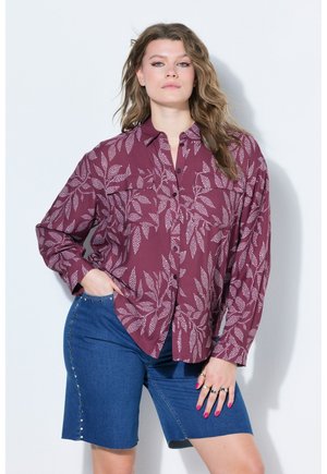 Kvinde iført en vinrød skjorte med knapper med hvide bladmønstre og blå denimshorts med metalnitter, stående med den ene hånd i lommen.