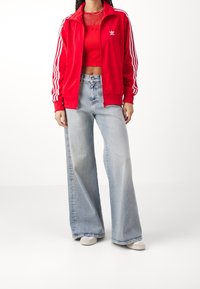 Czerwona kurtka na zamek z białymi paskami, zestawiona z czerwonym krótkim topem i jasnoniebieskimi szerokimi dżinsami. Na nogach białe trampki.
