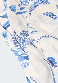 Chemise en coton avec un fond blanc ornée de motifs floraux et de feuilles bleus, fermeture à boutons et texture douce.