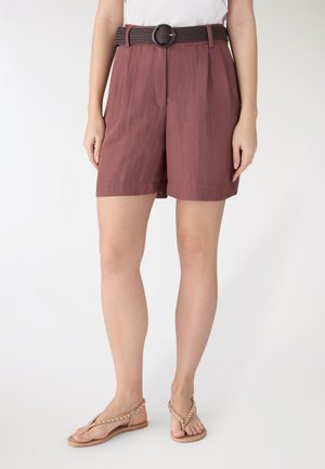 Donna che indossa pantaloncini mauve a vita alta con una cintura intrecciata marrone scuro e sandali piatti beige con borchie, in piedi davanti a uno sfondo neutro.