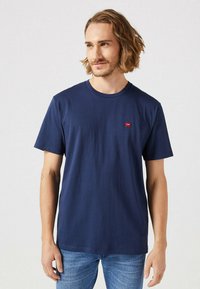 Navyblauwe katoenen t-shirt met korte mouwen, ronde hals en een klein rood logo-opdruk op de linkerborst. Zachte textuur, eenvoudig ontwerp.