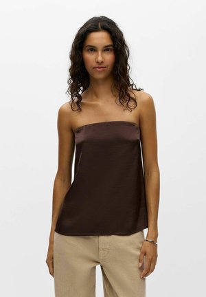 Femme aux cheveux bouclés foncés portant un haut en satin marron foncé sans manches et un pantalon beige, debout sur un fond blanc uni.