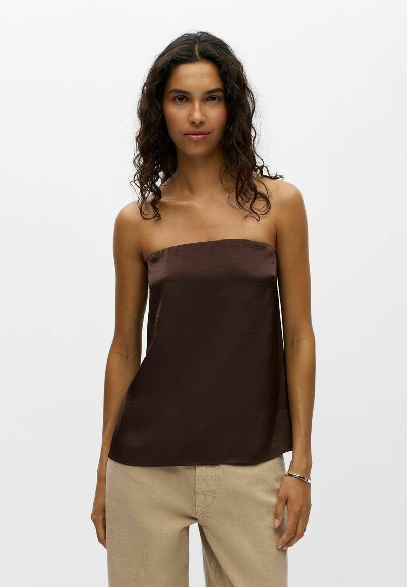 Femme aux cheveux bouclés foncés portant un haut en satin marron foncé sans manches et un pantalon beige, debout sur un fond blanc uni.