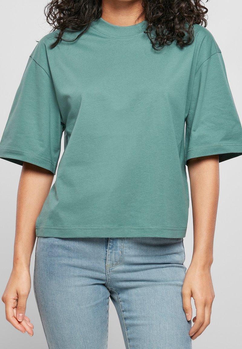 Femme portant un t-shirt vert ample à manches courtes et un jean bleu clair, debout devant un fond uni.