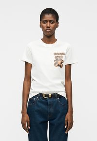 Mujer vistiendo una camiseta blanca con un gráfico de oso de peluche y texto "MOSCHINO", combinada con jeans azul oscuro y un cinturón negro.