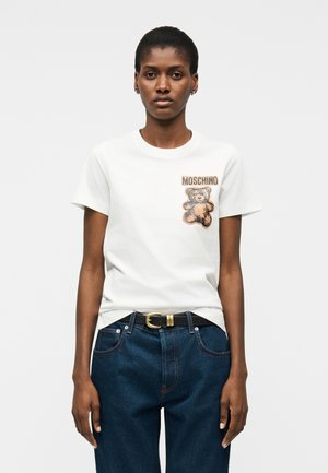 Γυναίκα που φοράει λευκό t-shirt με γραφικό αρκουδάκι και το κείμενο "MOSCHINO", συνδυασμένο με σκούρο μπλε τζιν και μαύρη ζώνη.