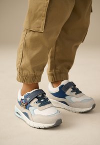 Børnesneakers med materialer i hvid, grå og blå. Har en frontrem, snørebånd og en tegneseriefigur som detalje på siden.