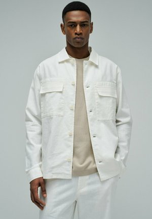 Homme portant une veste blanche boutonnée avec de grandes poches poitrine sur une chemise beige, debout avec une main dans la poche devant un fond uni.