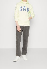 Neon gele hoodie met blauw "GAP" logo, aangesloten design en trekkoord. Gecombineerd met donkergrijze jeans en witte sneakers. Glad stoftextuur.
