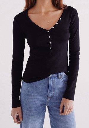 Femme portant un haut henley noir à manches longues côtelées avec des boutons blancs et un jean taille haute bleu clair, debout devant un fond uni.