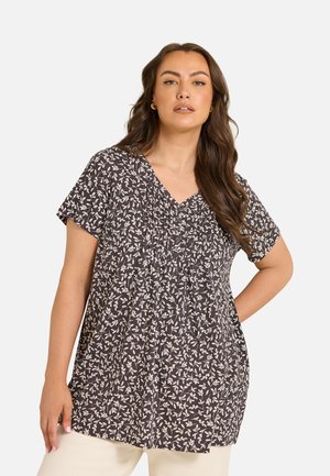 Mujer con el pelo largo y castaño lleva una blusa oscura de manga corta con un estampado floral blanco y pantalones claros, de pie contra un fondo liso.