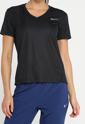 Femme portant un t-shirt noir à manches courtes et col en V de Nike, ainsi qu'un pantalon de sport bleu Nike, sur un fond blanc uni.