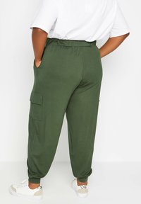Personne portant un pantalon cargo jogger vert olive avec une taille élastique et des poignets élastiques, des baskets blanches et un haut blanc à manches courtes, les mains dans les poches.