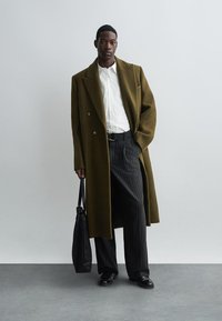 Abrigo de doble botonadura en verde oliva sobre camisa blanca, combinado con pantalones de rayas y zapatos negros, llevando un bolso tote negro.