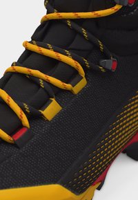 Μαύρο και κίτρινο παπούτσι πεζοπορίας με επενδυμένη γλώσσα, κορδόνια σε κίτρινο και μαύρο μοτίβο, και υφή σόλας με κόκκινες λεπτομέρειες.