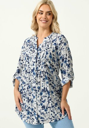 Donna che indossa una camicetta bianca con stampa floreale blu, maniche a tre quarti arrotolate, sorridente e in posa con le mani vicino ai fianchi.