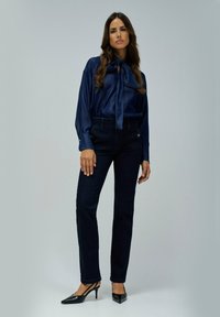 Blusa azul marino con cuello de lazo y puños abotonados, combinada con pantalones de mezclilla oscuros con bolsillos laterales y tacones negros puntiagudos.