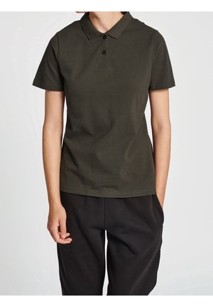 Persoon die een donkergroen poloshirt met korte mouwen en een zwarte broek draagt, staat tegen een egale lichte achtergrond.