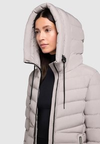 Doudoune gris clair avec une grande capuche, design matelassé, fermeture éclair noire et cordons de serrage pour ajustement. Textura lisse.