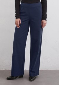 Pantaloni largi de culoare bleumarin, cu o textura netedă, talie ajustată și două pliuri frontale. Asortați cu pantofi negri cu toc.
